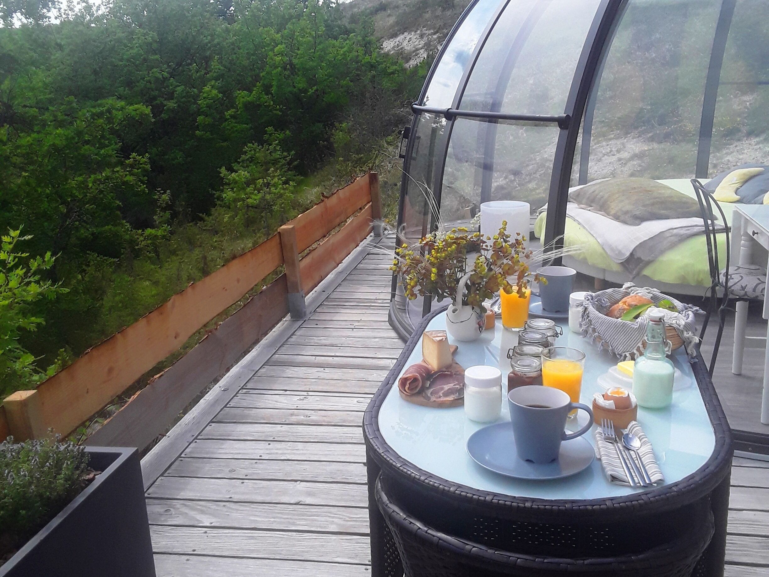 A breakfast table with food and drinks, a wicker chair, and a glass enclosure overlooking a forested area.