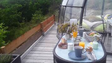 A breakfast table with food and drinks, a wicker chair, and a glass enclosure overlooking a forested area.