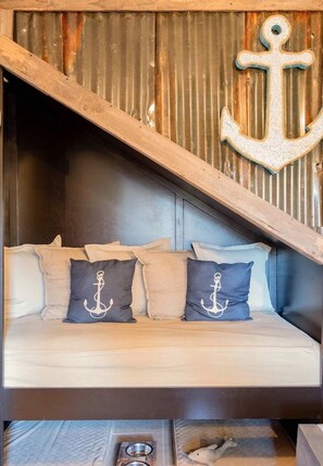 A cozy seating area with a wooden sloped ceiling, a metal wall with an anchor decoration, and a cushioned bench with pillows featuring anchor designs.