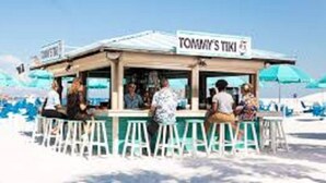 A beachside kiosk named "Tommy's Tiki" with a counter and stools, surrounded by beach chairs and umbrellas.