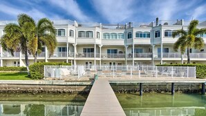 A waterfront hotel with a wooden pier leading to the building, featuring balconies and a reflection in the water.