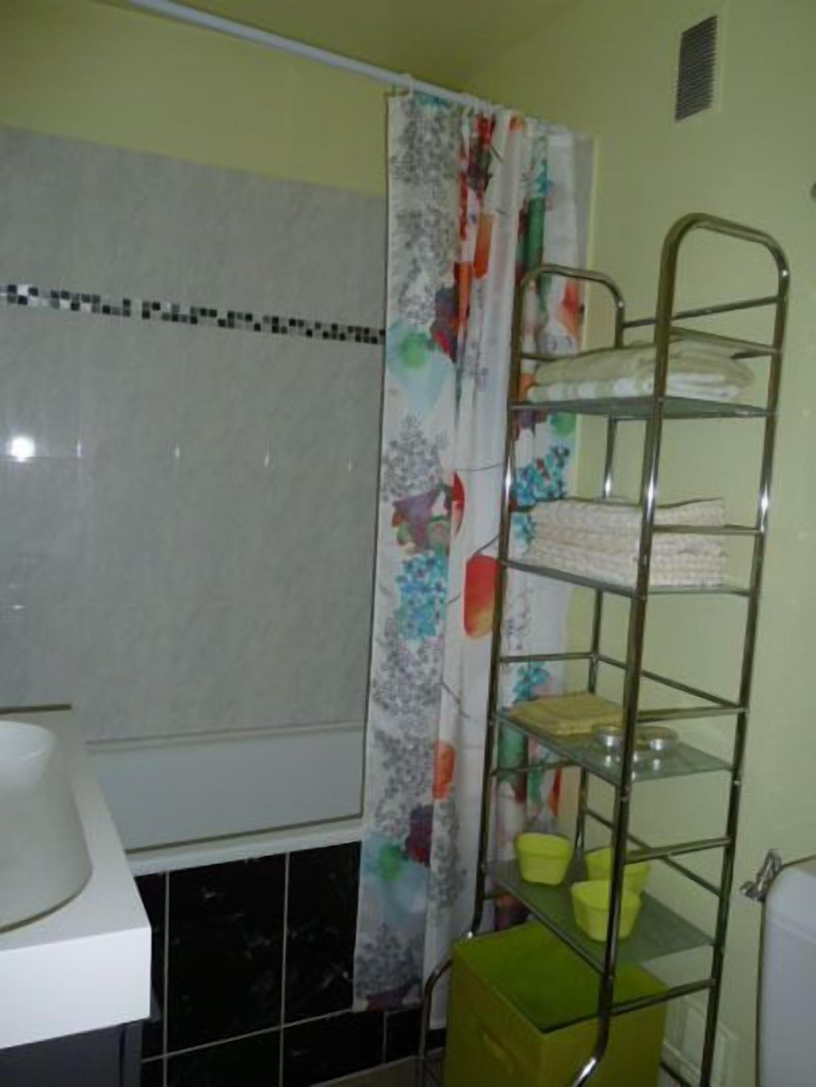 Un baño con inodoro, lavamanos, cortina de ducha con estampado floral y un estante metálico con recipientes amarillos.
