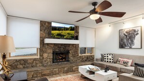 A modern living room with a stone fireplace, a ceiling fan, and a large TV mounted on the wall. - Tamarack - Steps to the Slopes + ASE Transportation (Snowmass Village)