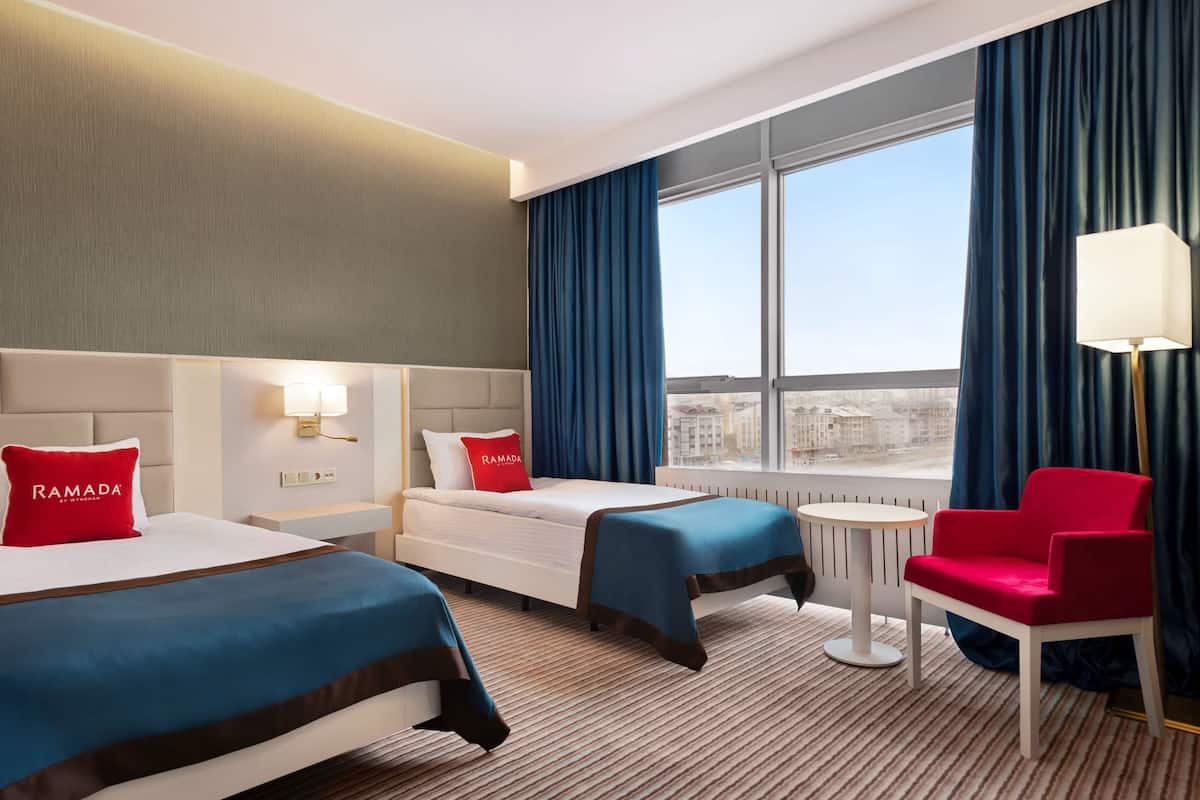Une chambre d’hôtel avec deux lits, un fauteuil rouge, une petite table et une vue sur la ville grâce à de grandes fenêtres.