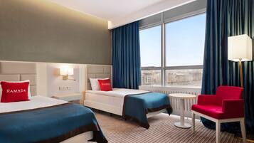 A hotel room with two beds, a red chair, a small table, and a view of the city through large windows.