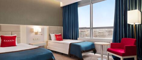 A hotel room with two beds, a red chair, a small table, and a view of the city through large windows.