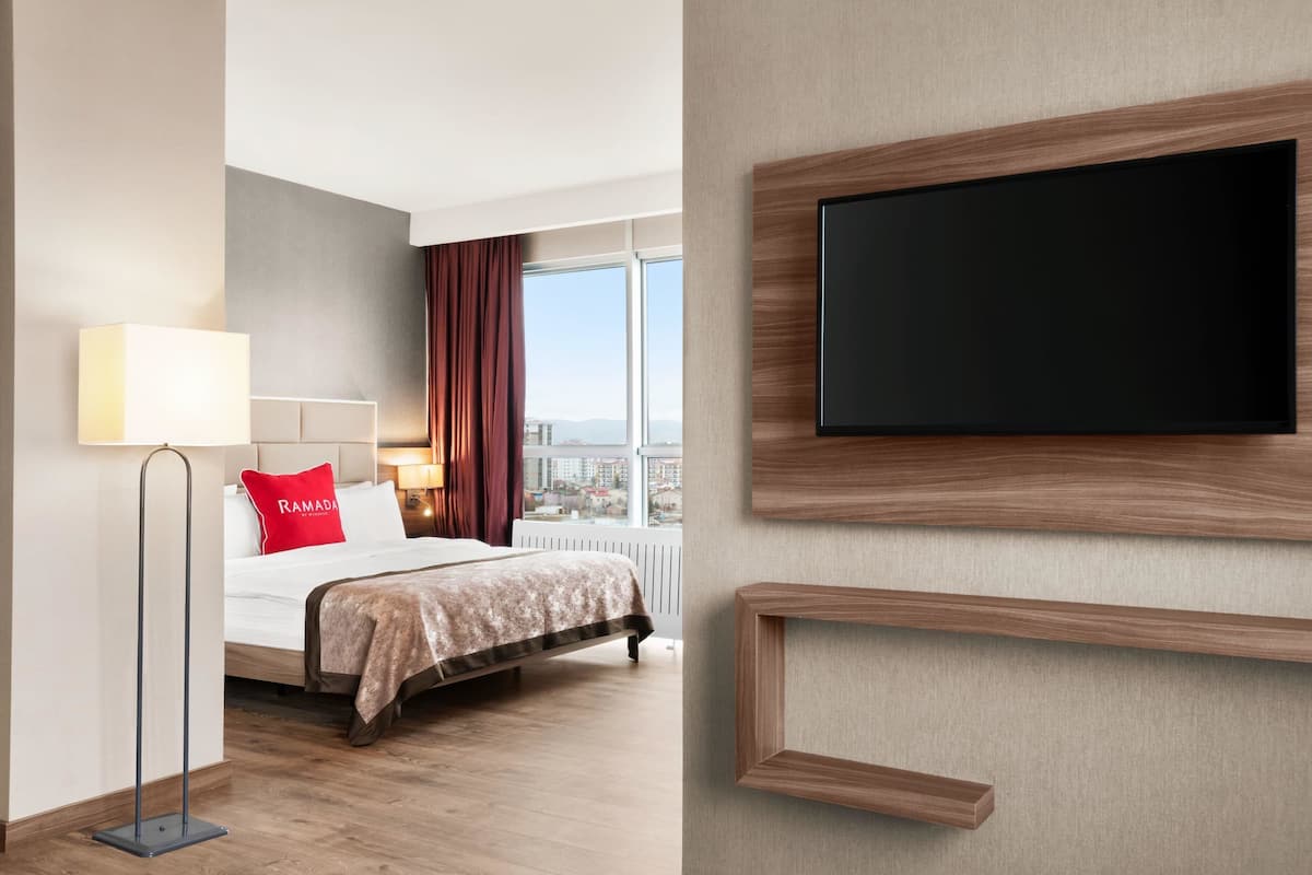 Une chambre d’hôtel avec un lit, un téléviseur fixé au mur, une lampe sur pied et une vue sur la ville grâce à de grandes fenêtres.