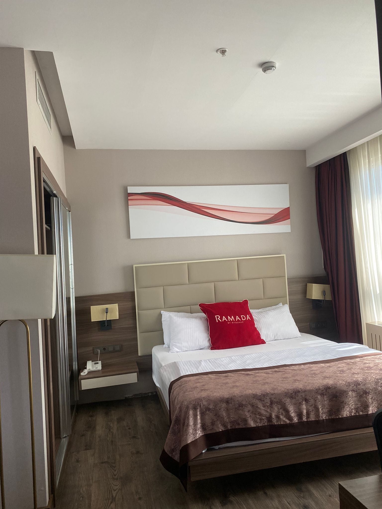 A hotel room with a bed, a nightstand, a lamp, a wall-mounted artwork, and a window with curtains.