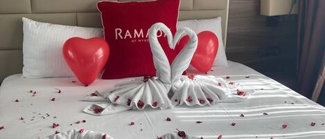 Una cama bien hecha con toallas con forma de corazón y pétalos de rosa, un cojín rojo con el texto 'RAM' y un globo rojo sobre una mesita de noche.