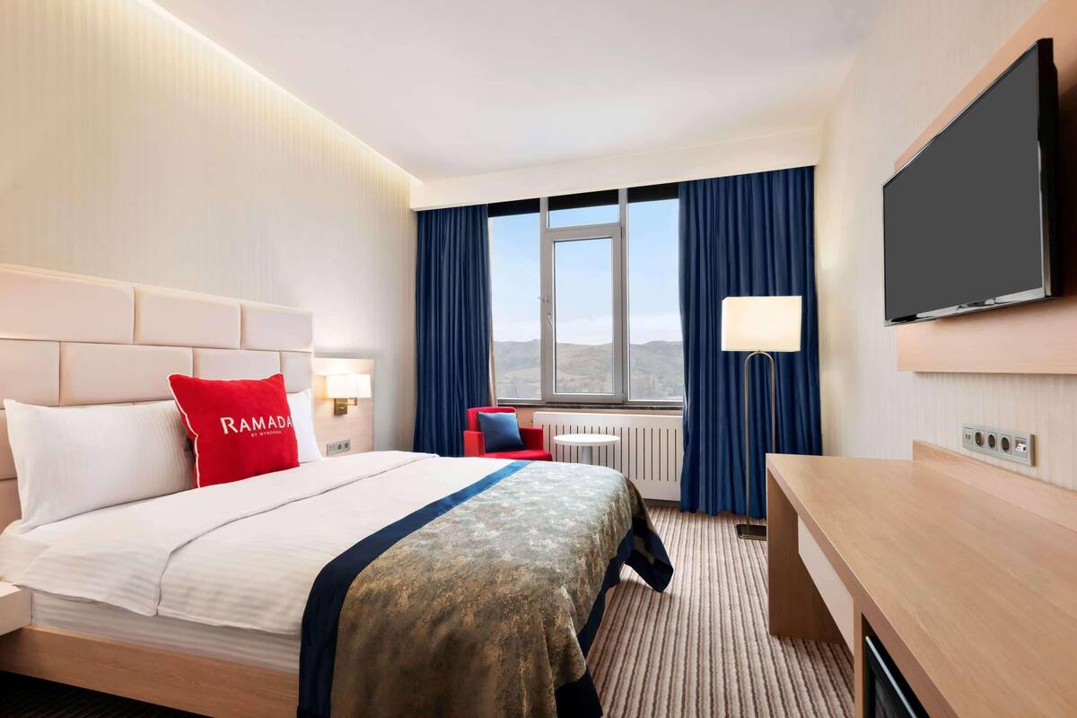 A hotel room with a bed, a desk, a TV, a window with curtains, and a view of the mountains.