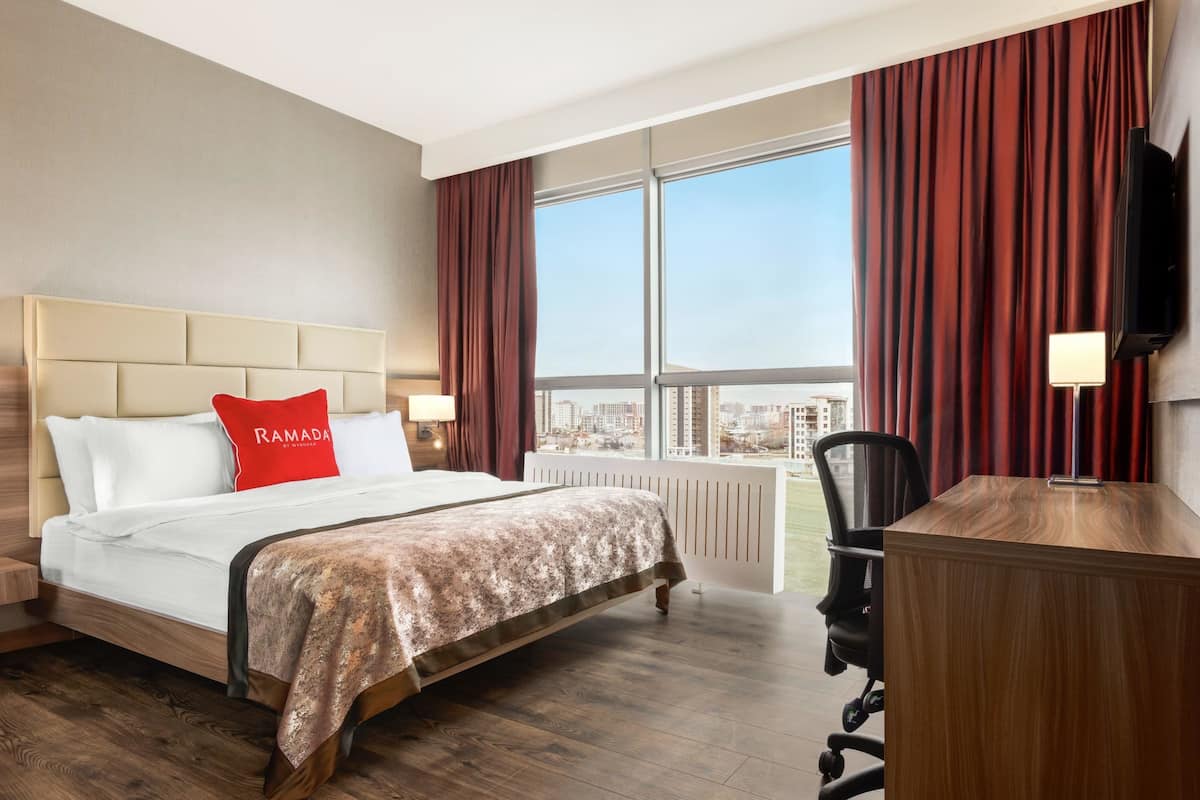 Une chambre d’hôtel avec un grand lit, un bureau, une chaise et une vue sur la ville grâce à de grandes fenêtres.