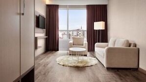 Una habitación de hotel moderna con un sofá, una pequeña mesa redonda, un televisor montado en la pared y vistas de la ciudad a través de amplios ventanales.