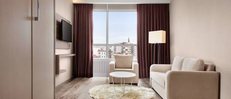 Una habitación de hotel moderna con un sofá, una pequeña mesa redonda, un televisor montado en la pared y vistas de la ciudad a través de amplios ventanales.