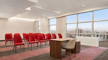 Un salón de conferencias con sillas rojas, una mesa de madera y ventanas amplias con vista a la ciudad.
