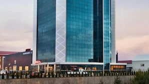 Un hotel moderno con fachada de vidrio, dos letreros destacados que dicen "RAMADA" en la parte superior, y un estacionamiento al frente.