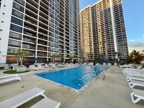 A swimming pool surrounded by lounge chairs, with high-rise buildings in the background.