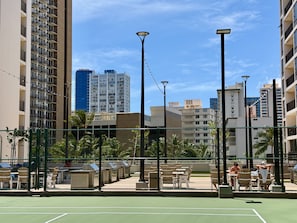 A tennis court with outdoor seating, surrounded by a chain-link fence, and high-rise buildings in the background.