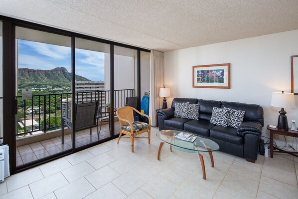 A modern living room with a glass coffee table, a black leather sofa with patterned cushions, a wooden chair, and a view of a mountainous landscape through large sliding doors.