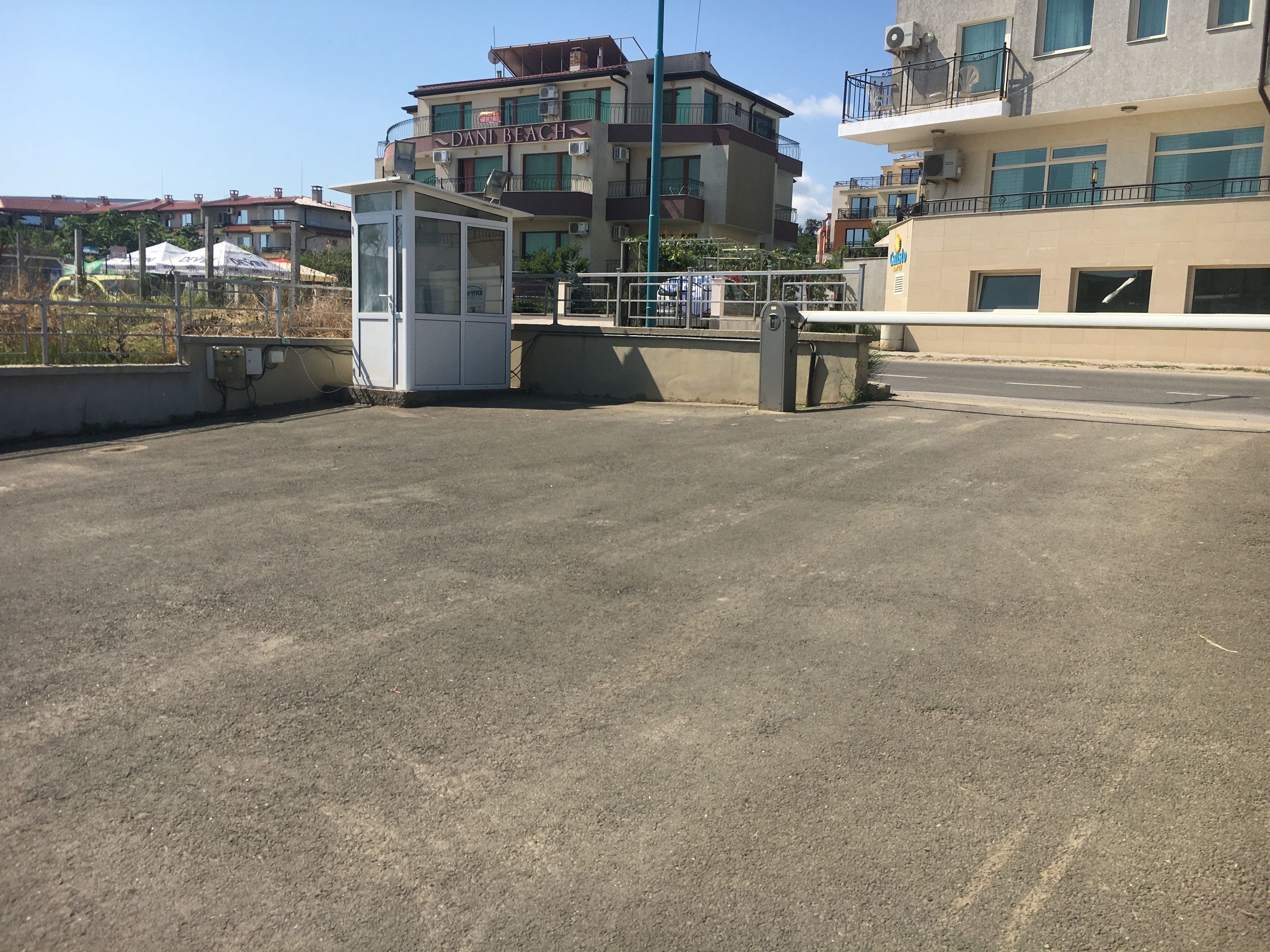 A parking area with a small kiosk, a building labeled "Dani Beach" with balconies, and a clear sky.