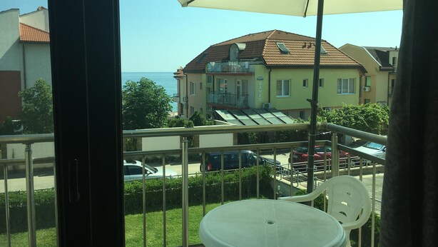 A balcony with a white table and chair, overlooking a street with parked cars and a building with a sign reading "Hotel Bar Rex." - Budzhaka Black Sea Paradise (Sozopol)