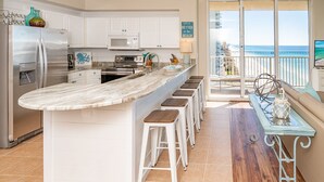 Une cuisine dotée d’un grand îlot central, de meubles blancs, d’appareils électroménagers en acier inoxydable et offrant une vue sur la plage.