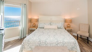 Une chambre avec un grand lit, deux tables de chevet avec des lampes, un fauteuil et une vue sur l’océan.