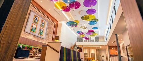 A modern hotel lobby with colorful hanging lamps, a staircase, and a seating area with a colorful striped wall.