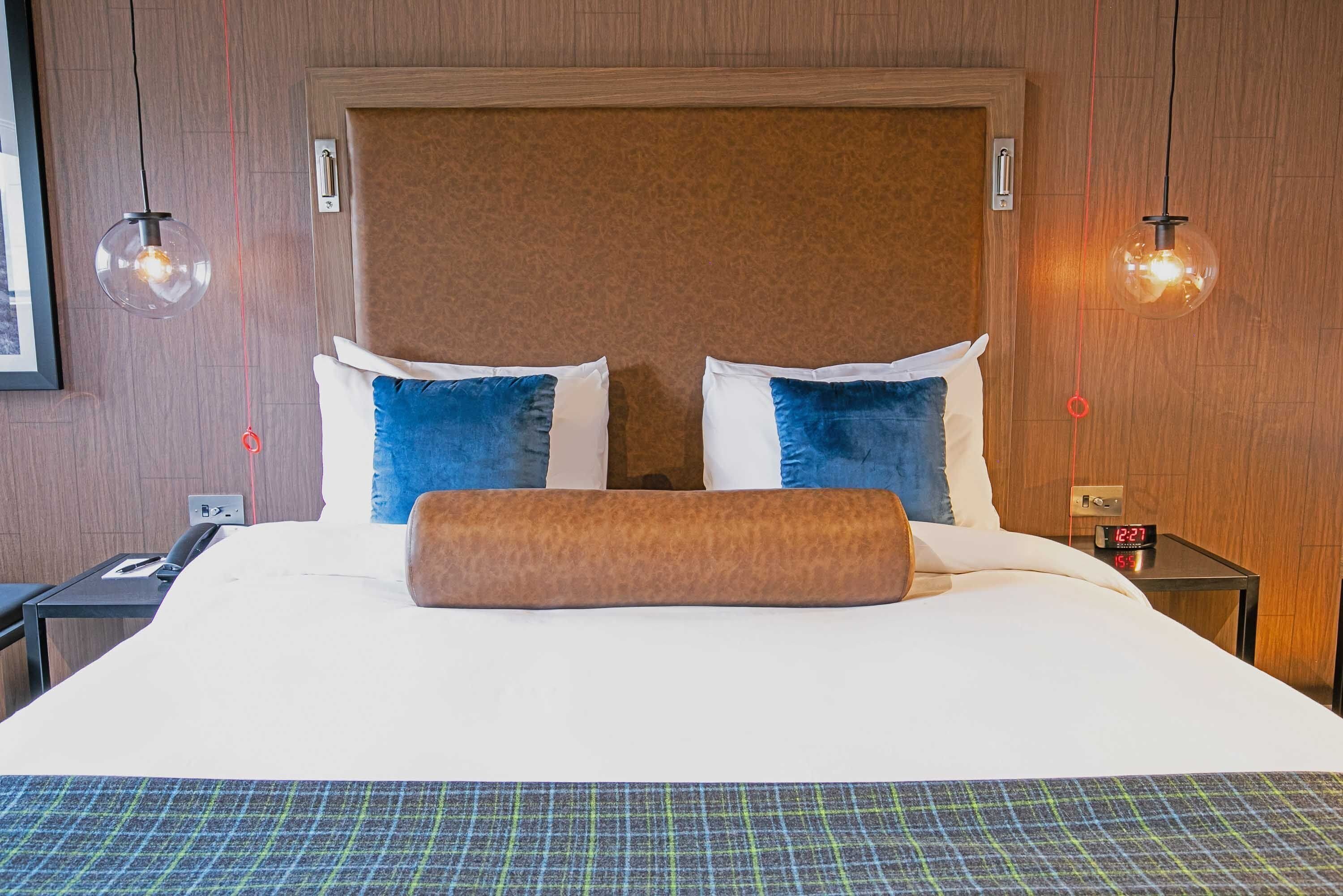 A hotel room with a large bed, two pillows, a blanket, a nightstand with a phone, and a wall clock.
