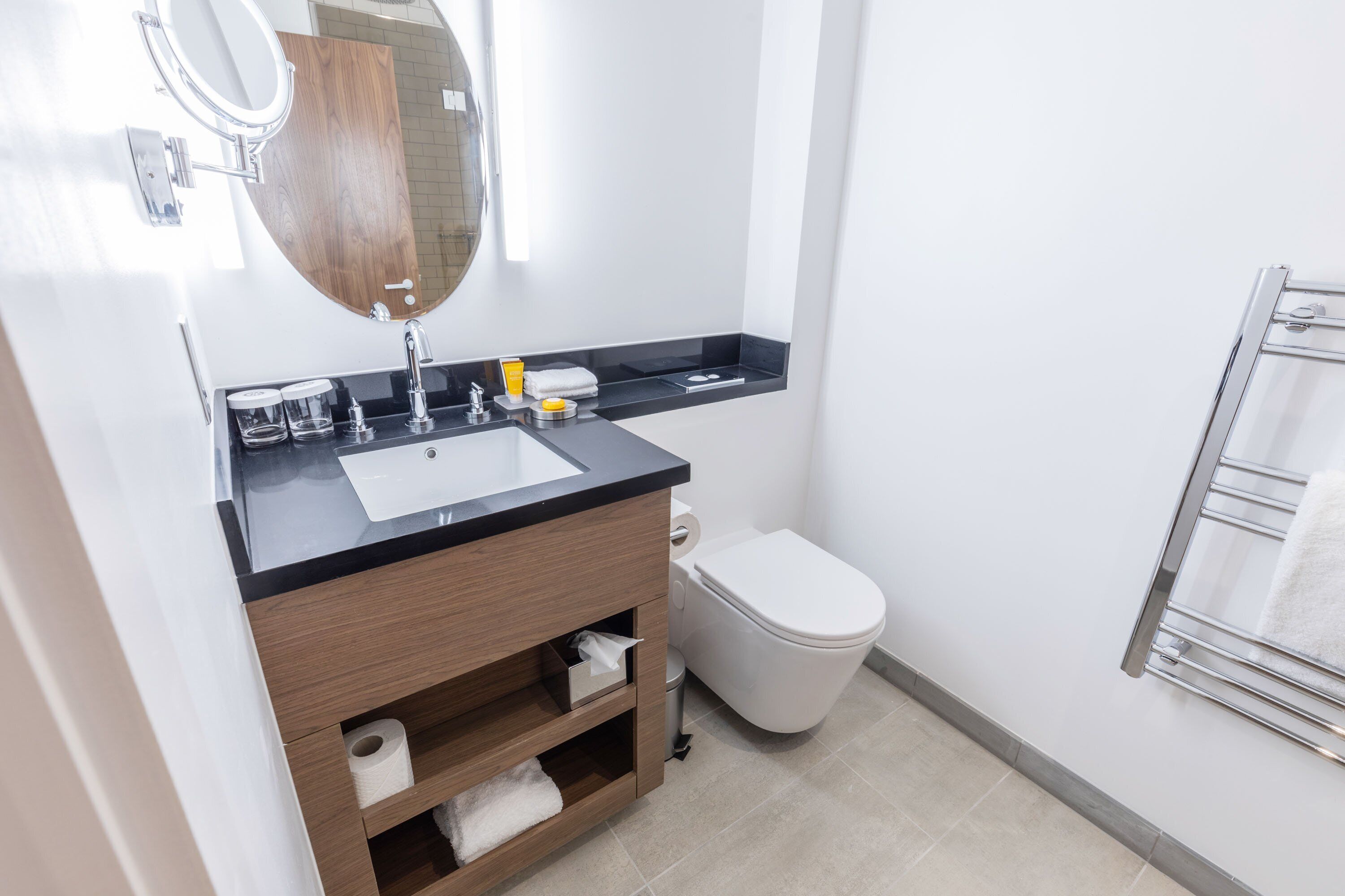 Un baño moderno con lavamanos blanco, tocador de madera, inodoro y perchero para toallas.