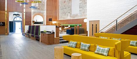 A modern hotel lobby with wooden floors, a yellow sofa, a reception desk, and a staircase.