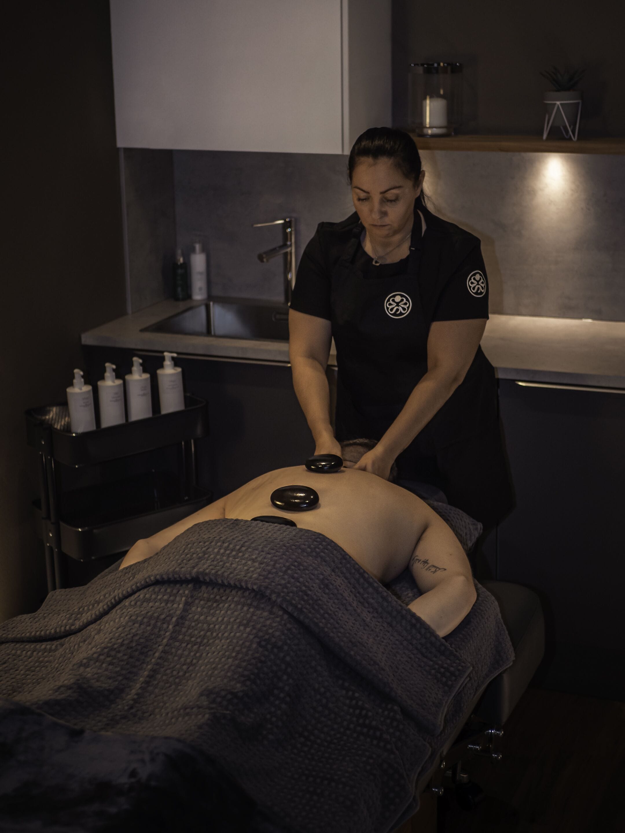A massage therapy session in a dimly lit room with a massage table, a sink, and a shelf with bottles.