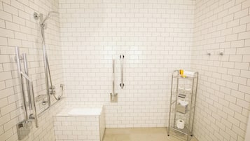 Une petite salle de bain aux murs recouverts de carreaux blancs, équipée d’une douche, de toilettes et d’une étagère en métal où sont rangés les articles de toilette.