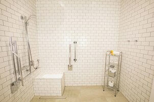 Une petite salle de bain aux murs recouverts de carreaux blancs, équipée d’une douche, de toilettes et d’une étagère en métal où sont rangés les articles de toilette.