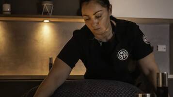 A massage therapist in a black uniform with a logo, working on a massage table with a pillow and a bottle of oil.
