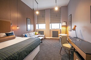 Quarto de hotel com uma cama grande, um sofá, uma escrivaninha e uma cadeira. Há duas janelas com persianas, um abajur e um quadro na parede.