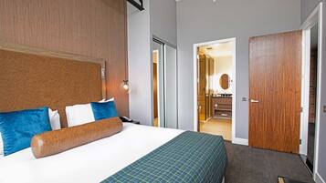 Una habitación de hotel con una cama grande, un cabecero de madera, una mesita de noche y una mesita auxiliar. Hay una puerta que da a un baño y un pasillo.