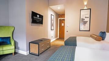 A hotel room with a green armchair, a bed with blue and white bedding, a small wooden cabinet, and a framed picture on the wall.