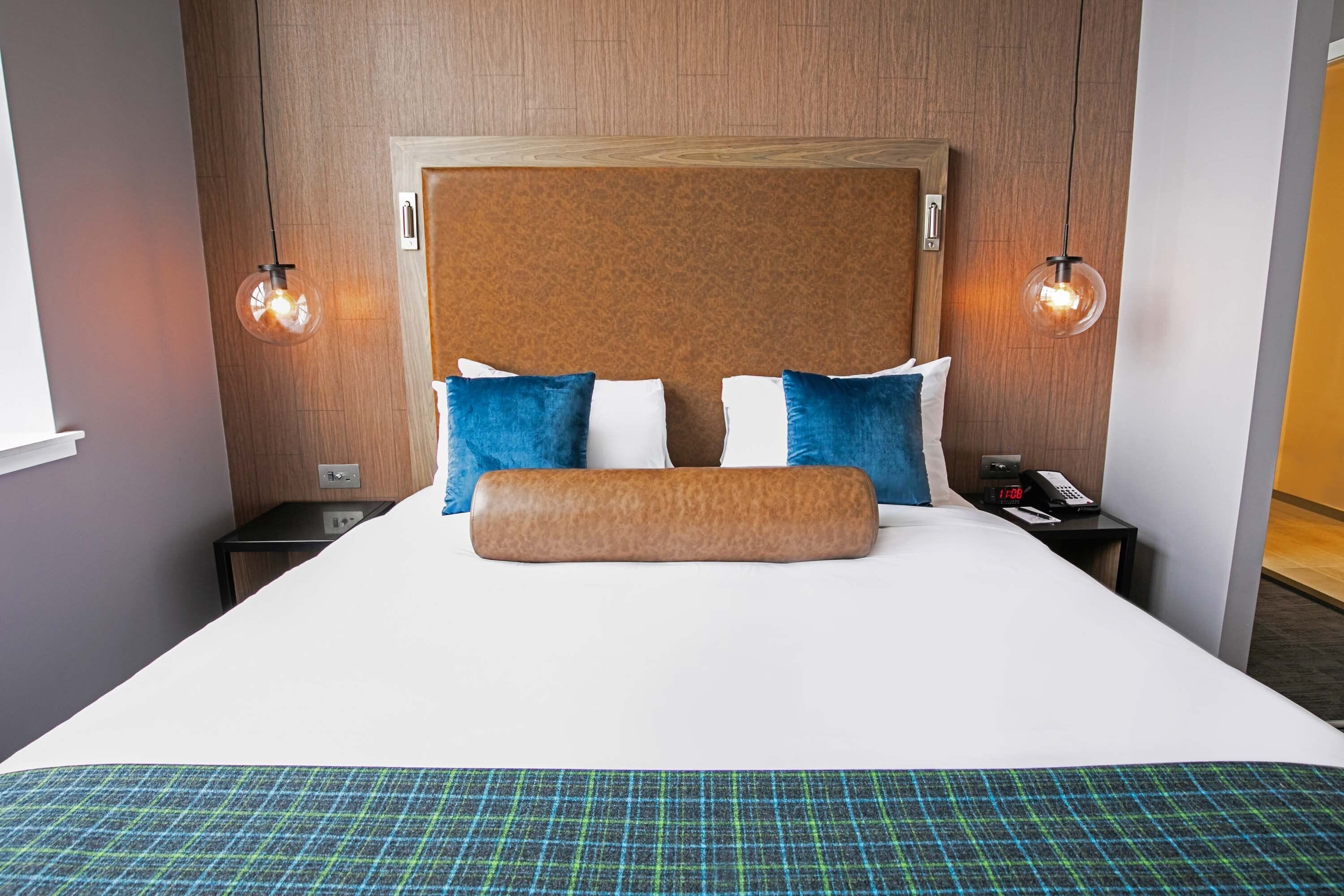 A hotel room with a large bed, two bedside tables, a wooden headboard, and two wall-mounted lamps.