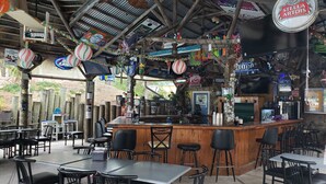 Un espace bar extérieur avec des tables et des chaises en bois, un comptoir avec une caisse enregistreuse, et diverses décorations, notamment des panneaux et des ballons.