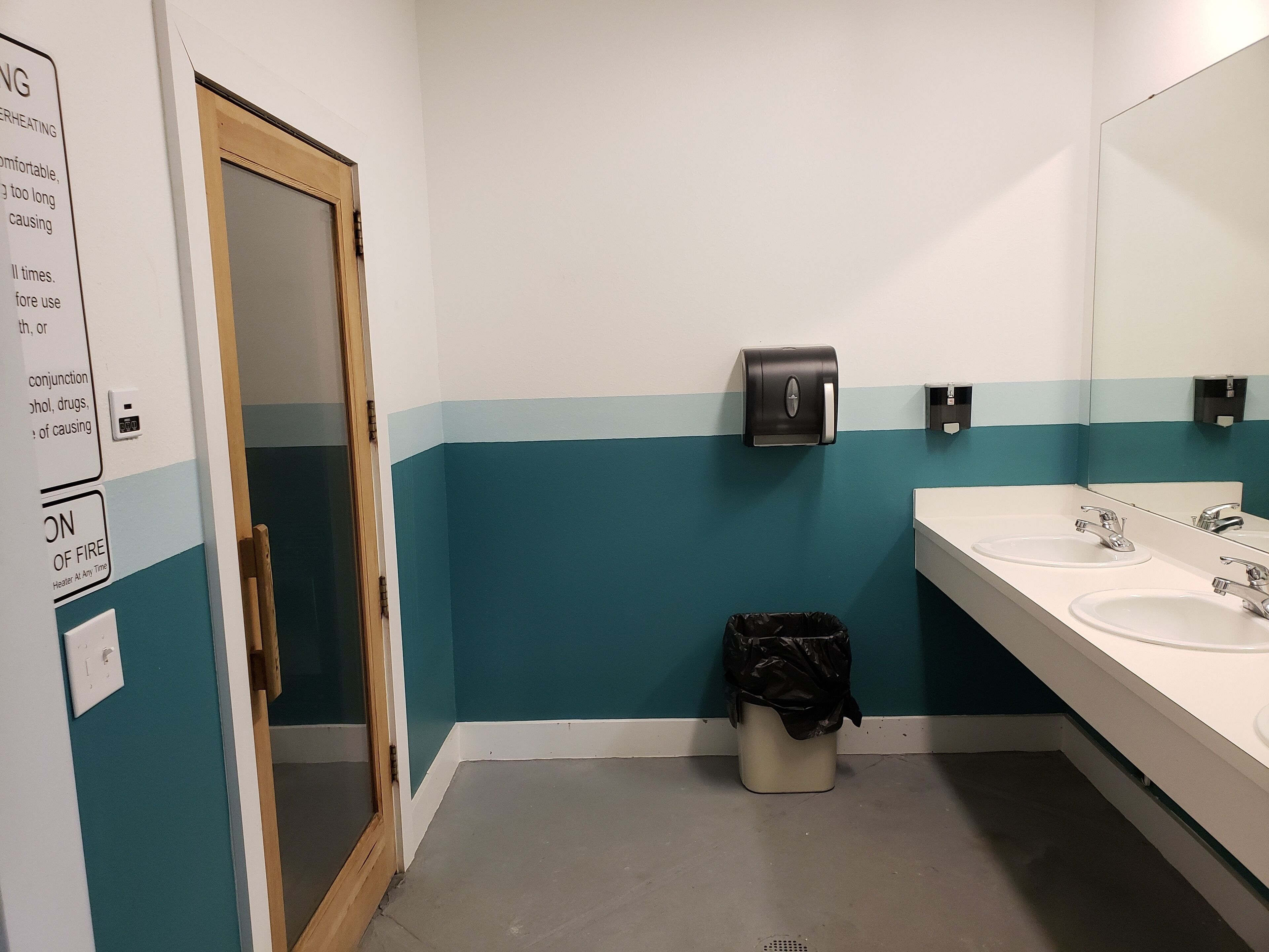A bathroom with a teal and white color scheme, a hand dryer, a trash bin, and a mirror above the sinks.