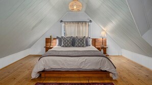 Ein Schlafzimmer mit schrägen Wänden, einem Bett mit grau-weißer Decke, gemusterten Kissen, Holz-Nachttischen und einer Pendelleuchte.