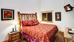 Un dormitorio con cama, cabecera de madera, mesita de noche, lámpara, cuadros enmarcados y televisor.
