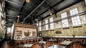 A spacious bar area with high ceilings, exposed beams, and a brick wall. There are multiple tables and chairs arranged for dining.
