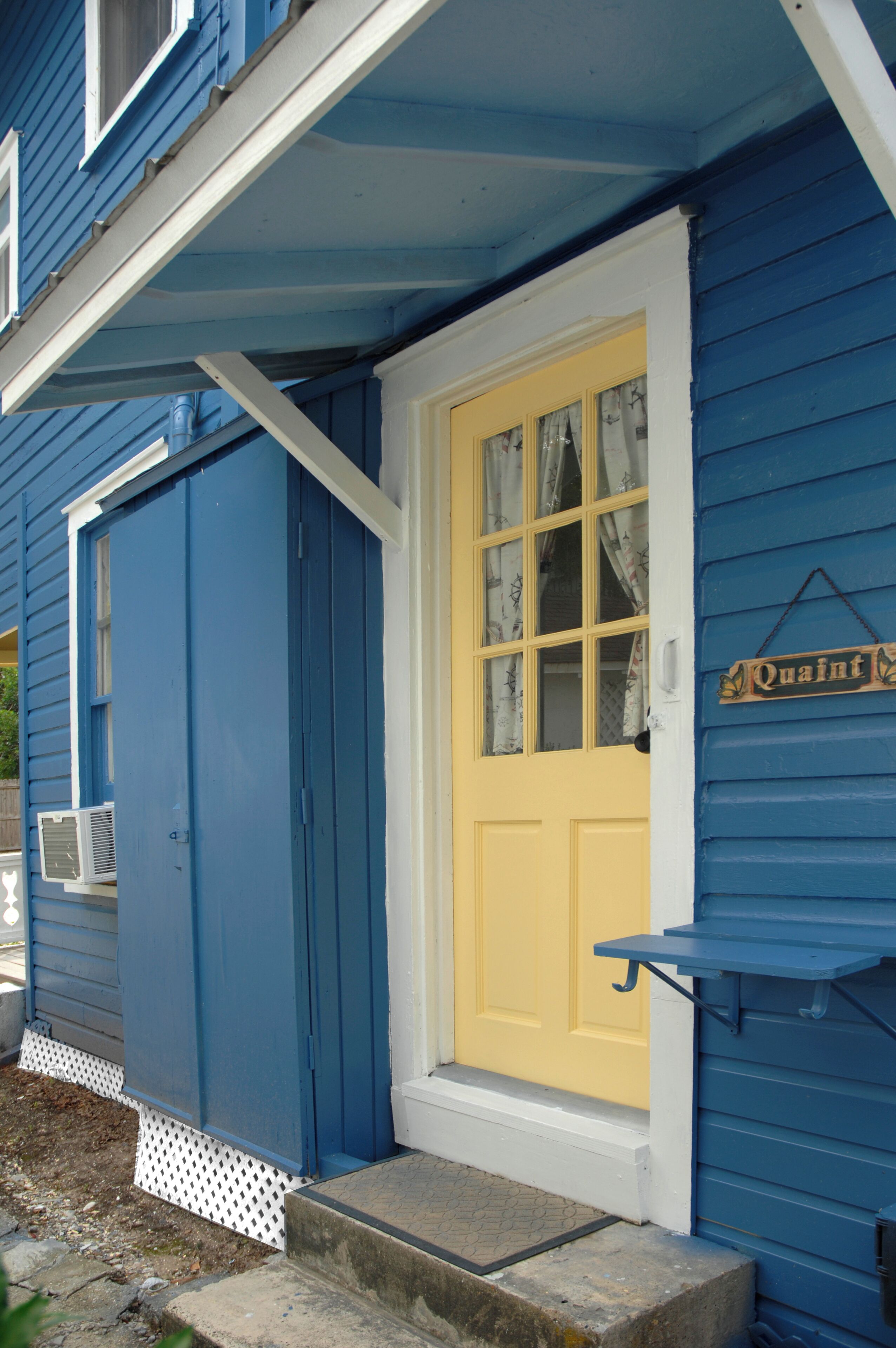 A blue house with a yellow door, a small porch, and a sign that reads "Quaint."