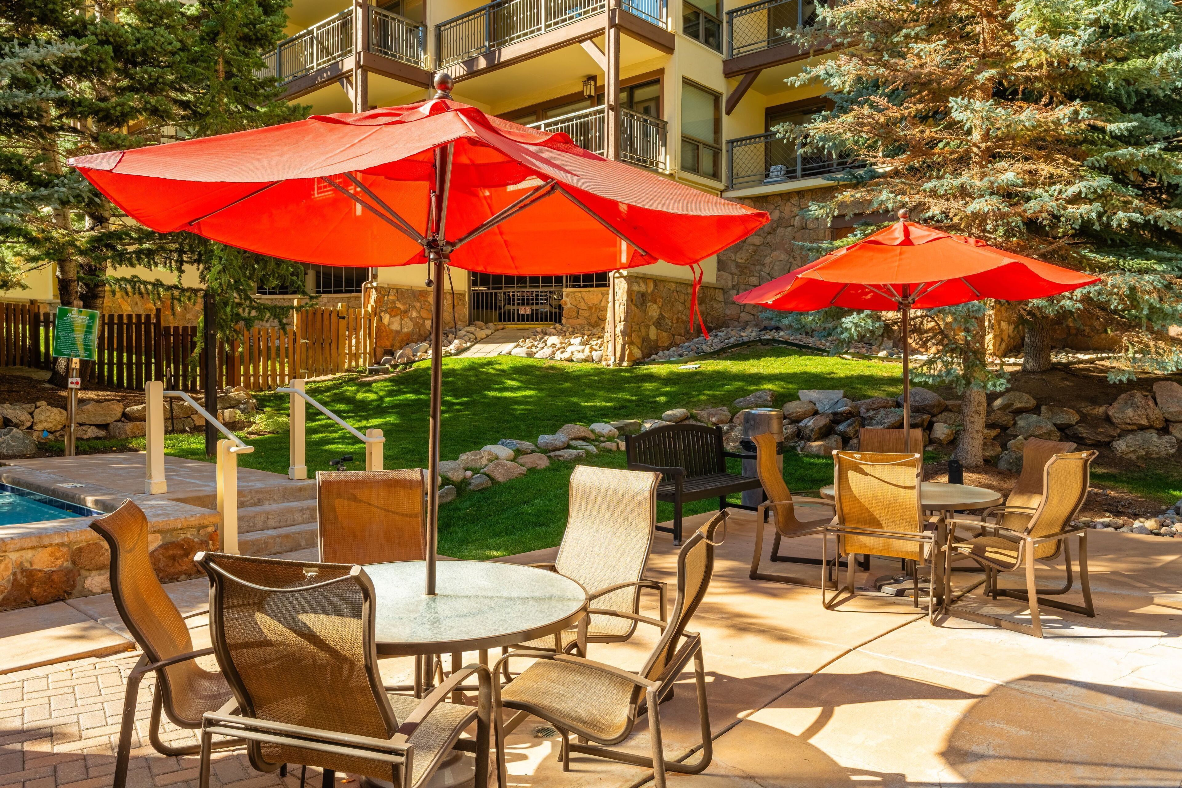 An outdoor patio area with round tables and chairs, red umbrellas, and a pool.