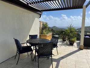 A covered outdoor seating area with a table and chairs, potted plants, and a view of the sea and hills. - Private pool 1-bed close to Cruz Bay Ocean views (St john)