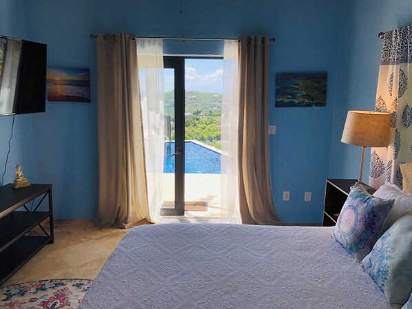 Habitación de hotel con cama, televisor, escritorio, lámpara y vistas a una piscina y zonas verdes.