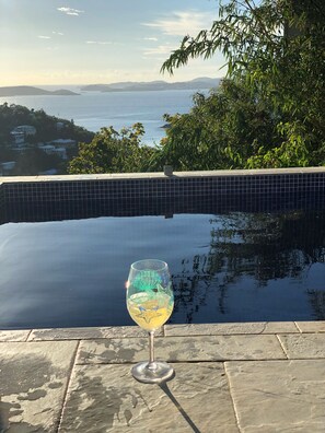 Un vaso de bebida sobre una repisa de piedra junto a una piscina, con vista a la costa distante.