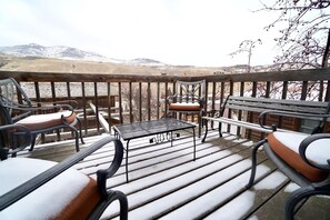 A wooden deck with a metal table and chairs, overlooking a snowy landscape with mountains.