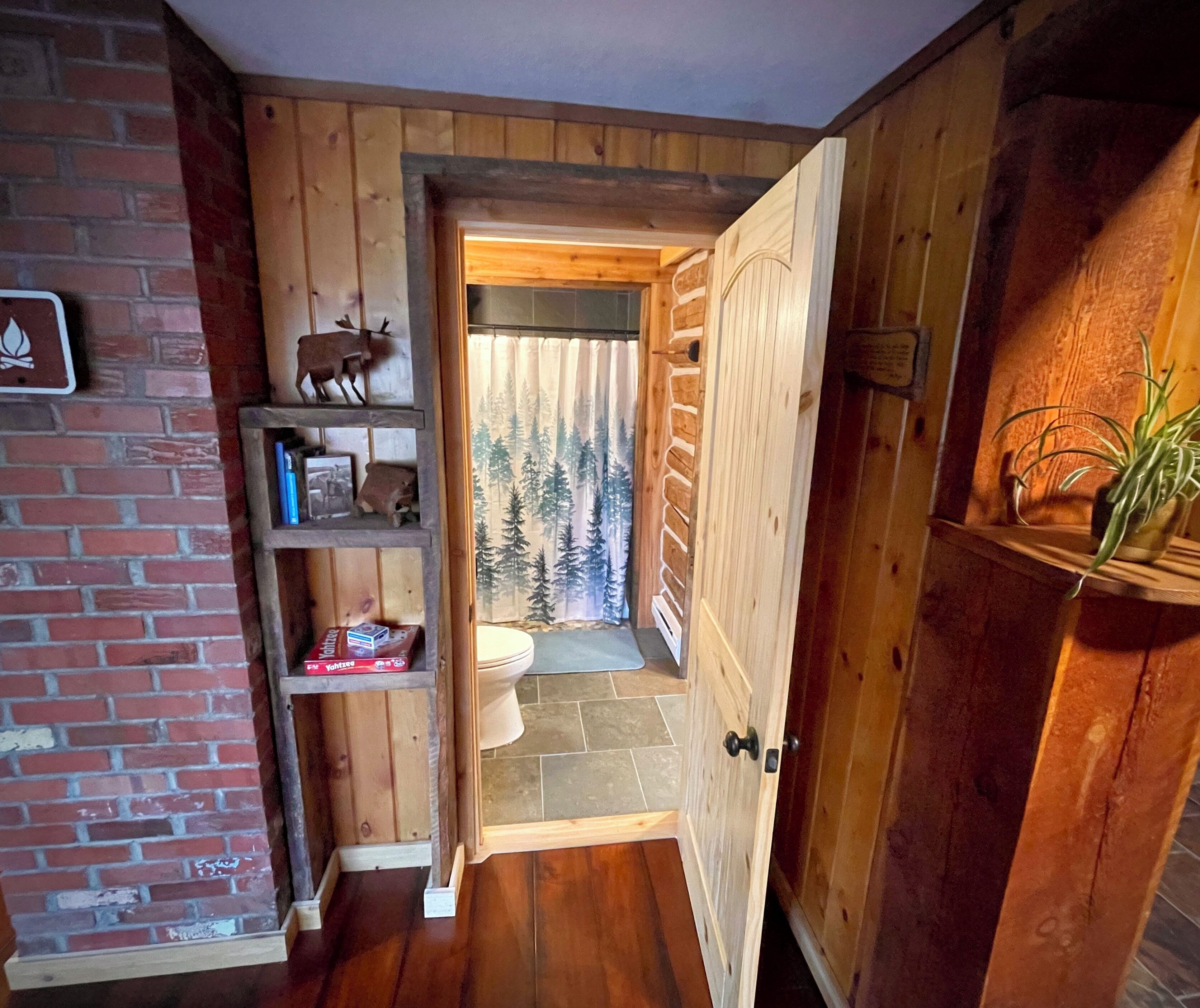 Un intérieur en bois avec une cheminée, une étagère remplie de livres et une porte donnant sur une salle de bain dont le rideau de douche est décoré de motifs forestiers.